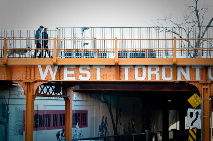 Two people walk a dog across an orange bridge with large white WEST TORONTO lettering.